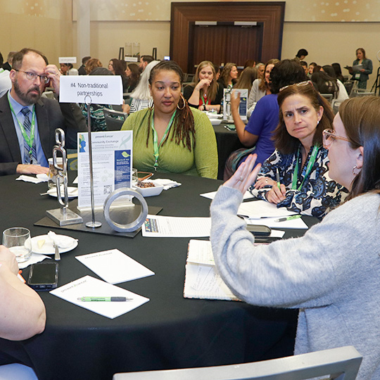 Groups of people sit at large, circular tables discussing topics.
