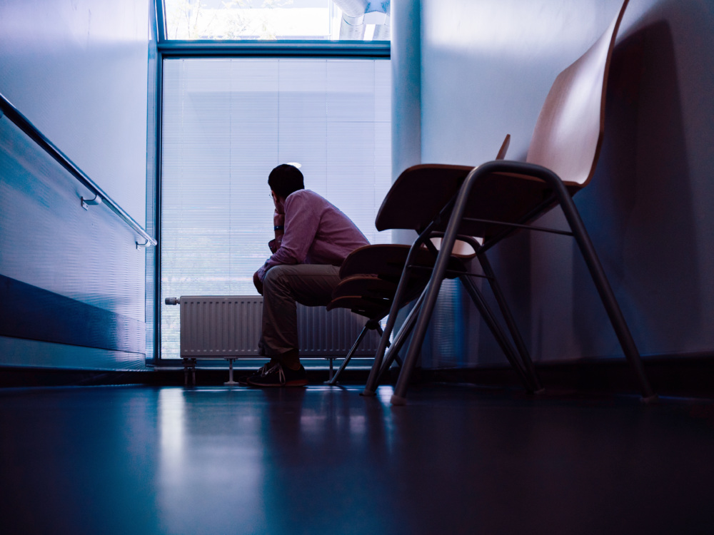 Low angle view of lonely patient sitting in modern hospital waiting lobby room looking through window with blinds as he waits for good or bad news from his doctor