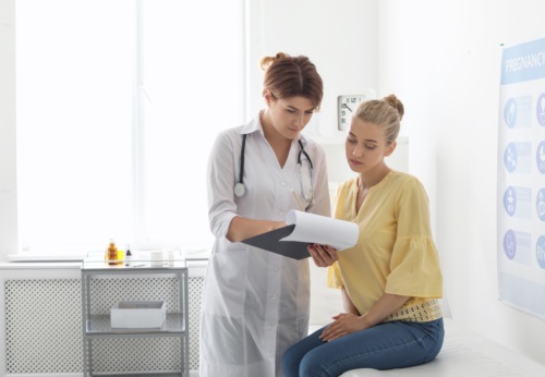 Female patient sitting on doctor's table chatting with female doctor looking at a clipboard.