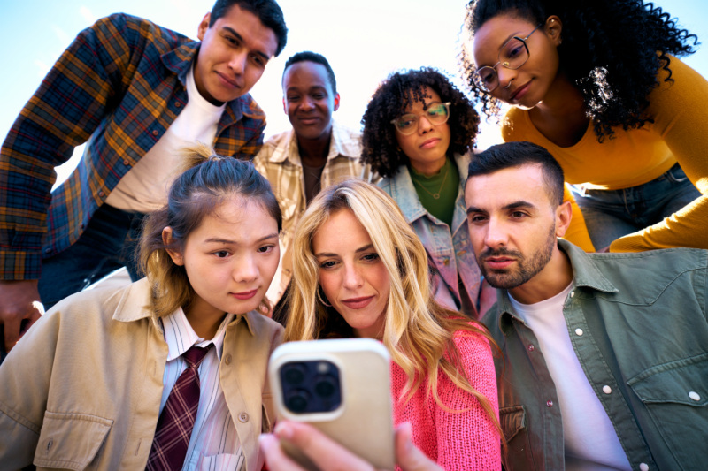 A group of twenty-year-olds all look at one phone with varying facial expressions.