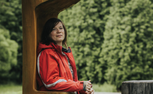 Portrait of a white woman in a red parka leaning against a cabin wall surrounded by green pine trees.