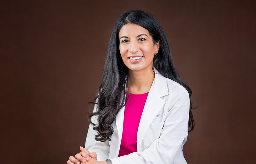 A woman with long, dark hair smiles while wearing a white lab coat and a pink shirt.